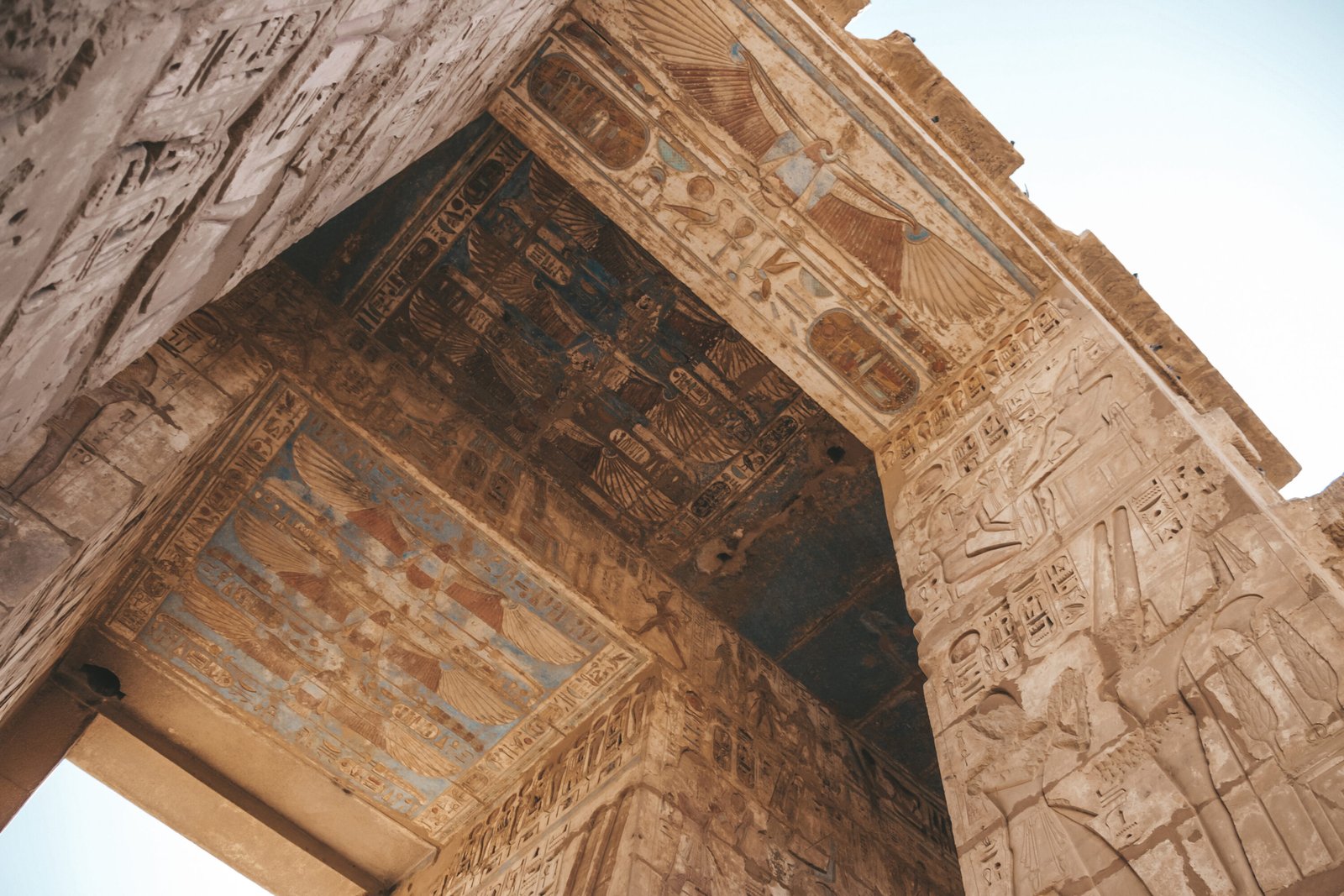 Upward view through a Karnak Temple gateway preserving blue, red and gold-painted winged vultures, royal cartouches, hieroglyphs and carved pharaonic reliefs