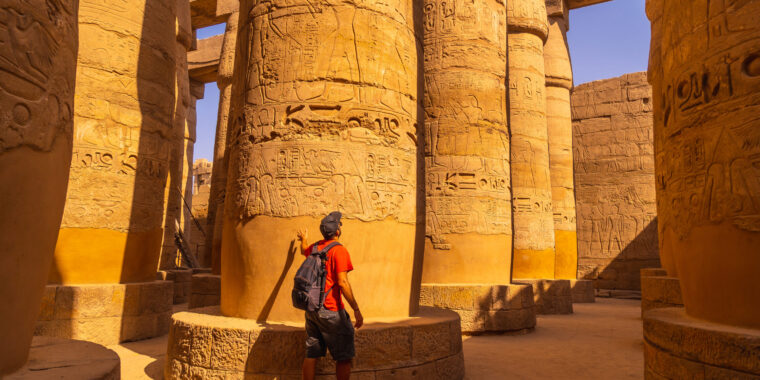 Visitor examining massive papyriform columns covered with Ramesside royal cartouches, offering scenes and hieroglyphs in Karnak’s Great Hypostyle Hall at Luxor
