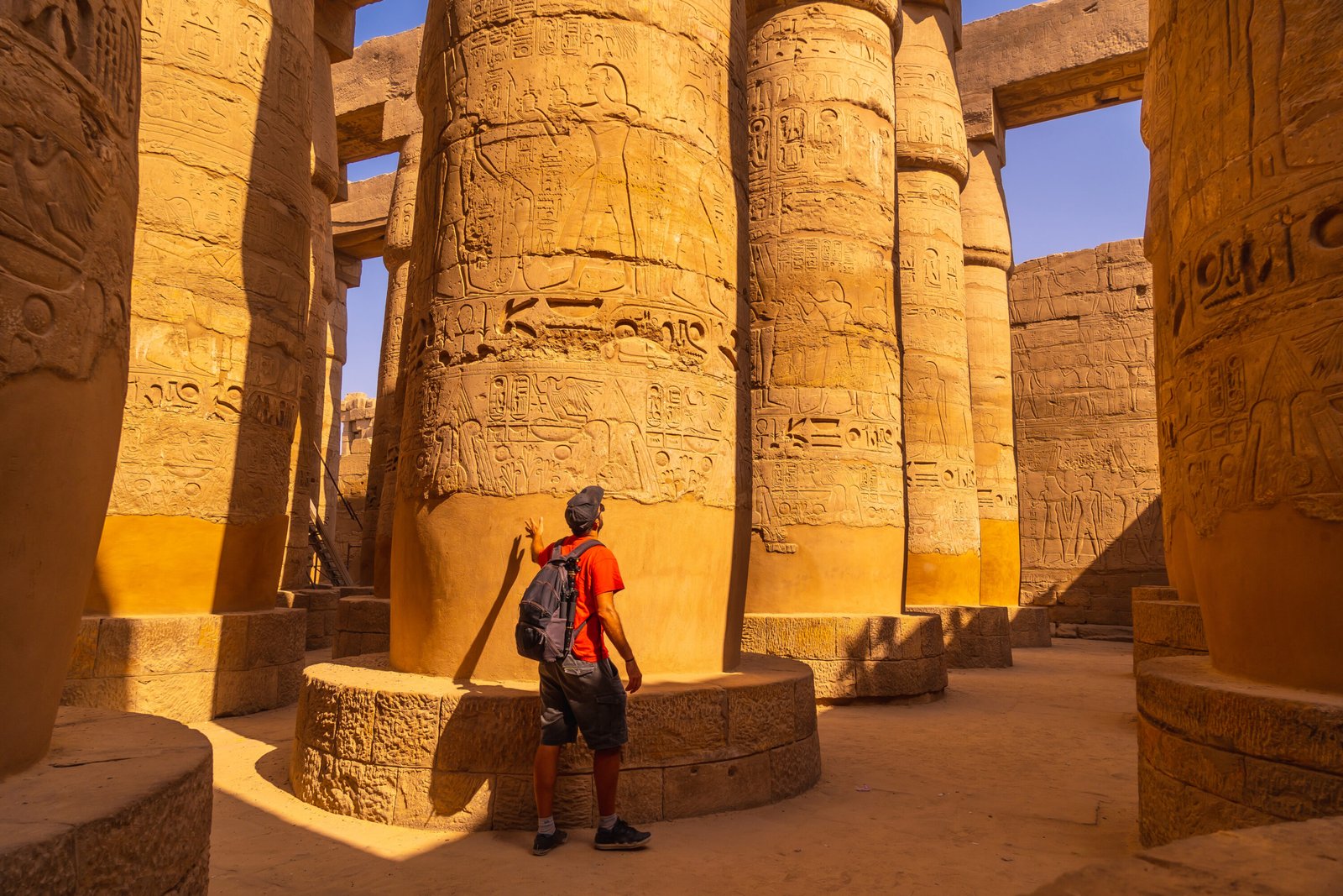 Visitor examining massive papyriform columns covered with Ramesside royal cartouches, offering scenes and hieroglyphs in Karnak’s Great Hypostyle Hall at Luxor