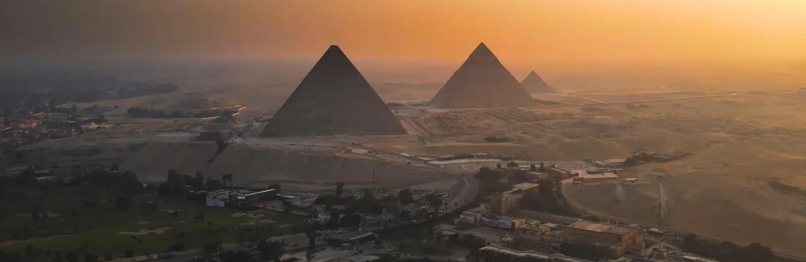 Elevated sunset view across the Giza Plateau showing the pyramids of Khufu, Khafre and Menkaure silhouetted beneath a glowing orange sky