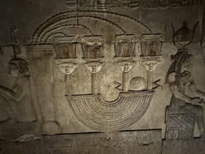 Dendera Temple relief showing four Hathor-headed sistrum emblems rising from an ornate ritual object, flanked by standing deities and hieroglyphic inscriptions