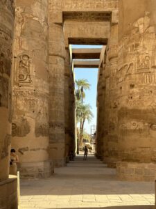 Long passage through the Great Hypostyle Hall at Karnak framed by hieroglyph-covered columns and painted architraves, opening toward sunlit palm trees outside
