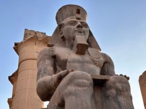 Low-angle view of a colossal seated statue of Pharaoh Ramesses II wearing the double crown and ceremonial beard in his first courtyard at Luxor Temple, Luxor, Egypt