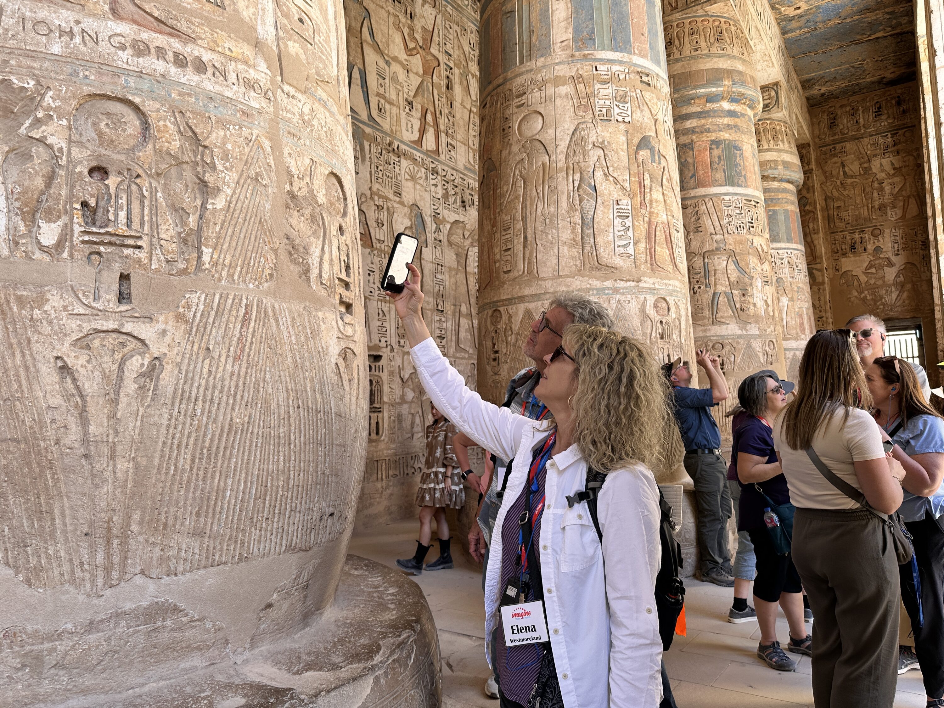 Egypt Christian Tours group examining painted and carved Ptolemaic columns inside the Temple of Horus at Edfu, with surviving blue, red and yellow pigment on reliefs and hieroglyphs