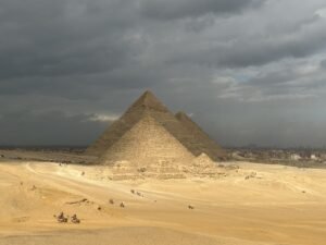 Pyramids rising from the Giza Plateau beneath dark storm clouds with visitors and camels below