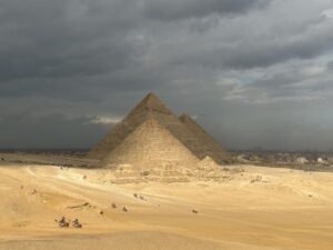 Pyramid of Menkaure in the foreground with Khafre and Khufu aligned behind it on the Giza Plateau beneath dark storm clouds, with camels and visitors crossing the desert