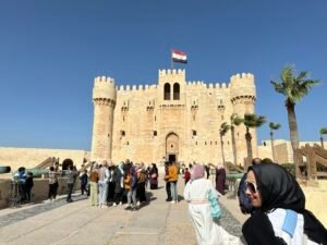 Stone fortress of the Citadel of Qaitbay in Alexandria, Egypt, with visitors, palm trees, and an Egyptian flag