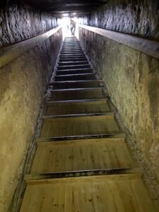 Steep wooden stairway climbing through a narrow stone passage toward a bright opening in an ancient Egyptian monument