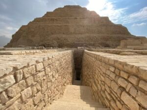 Ancient stone stairway descending between masonry walls with the Step Pyramid of Djoser rising behind at Saqqara