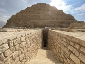 Ancient stone stairway descending between masonry walls with the Step Pyramid of Djoser rising behind at Saqqara