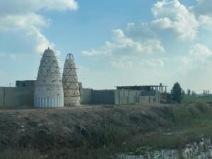 Two conical pigeon towers standing beside low buildings and farmland in rural Egypt beneath large clouds