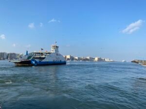 Blue-and-white passenger ferry crossing Mediterranean waters off Alexandria, Egypt, with the city shoreline behind