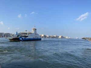 Blue-and-white passenger ferry crossing Mediterranean waters off Port Said, Egypt, with the city shoreline behind