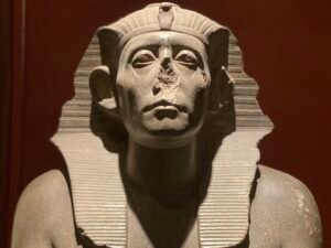 Dark stone ancient Egyptian royal statue with striped nemes headdress, uraeus and damaged nose displayed in the Egyptian Museum in Cairo