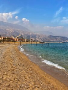 Pebbled Red Sea shoreline with clear blue water, small waves, and rugged mountains behind coastal buildings