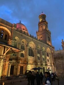 Historic Cairo mosque illuminated at dusk with a domed roof, tall minaret, and arched windows
