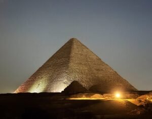 Great Pyramid of Khufu illuminated by warm spotlights at night with smaller pyramid structures silhouetted in the foreground