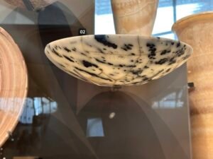 Shallow Predynastic Egyptian stone bowl with dark natural speckling and a finely polished rim, displayed among Naqada-period grave goods at the Grand Egyptian Museum in Giza
