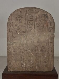 Round-topped Egyptian stone stela with a seated deity facing a standing worshipper above a lower register of family figures, with hieroglyphs and traces of red pigment, Egyptian Museum Cairo