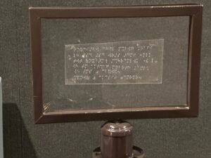 Framed transparent museum label with raised Braille text mounted above an exhibit stand
