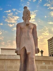 Tall ancient Egyptian pharaoh statue with crown and ceremonial beard silhouetted against a warm sunset sky
