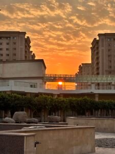 Orange sunset glowing between modern Cairo buildings beneath a textured cloud-filled sky