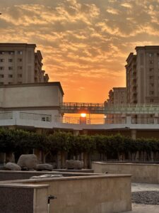 Orange sunset glowing between modern Cairo buildings beneath a textured cloud-filled sky