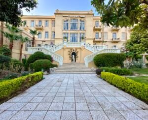 Historic Winter Palace hotel in Luxor viewed across a formal garden with trimmed hedges, palms, and a grand double staircase