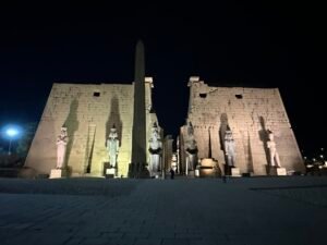 First pylon of Ramesses II at Luxor Temple illuminated at night with the surviving 25-meter obelisk and colossal seated royal statues at the temple entrance in Luxor, Egypt